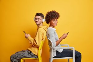 gadget addiction and lifestyle concept. relaxed pleased interracial couple sit back to each other ignore live communication use modern mobile phones pose on chairs against yellow background.
